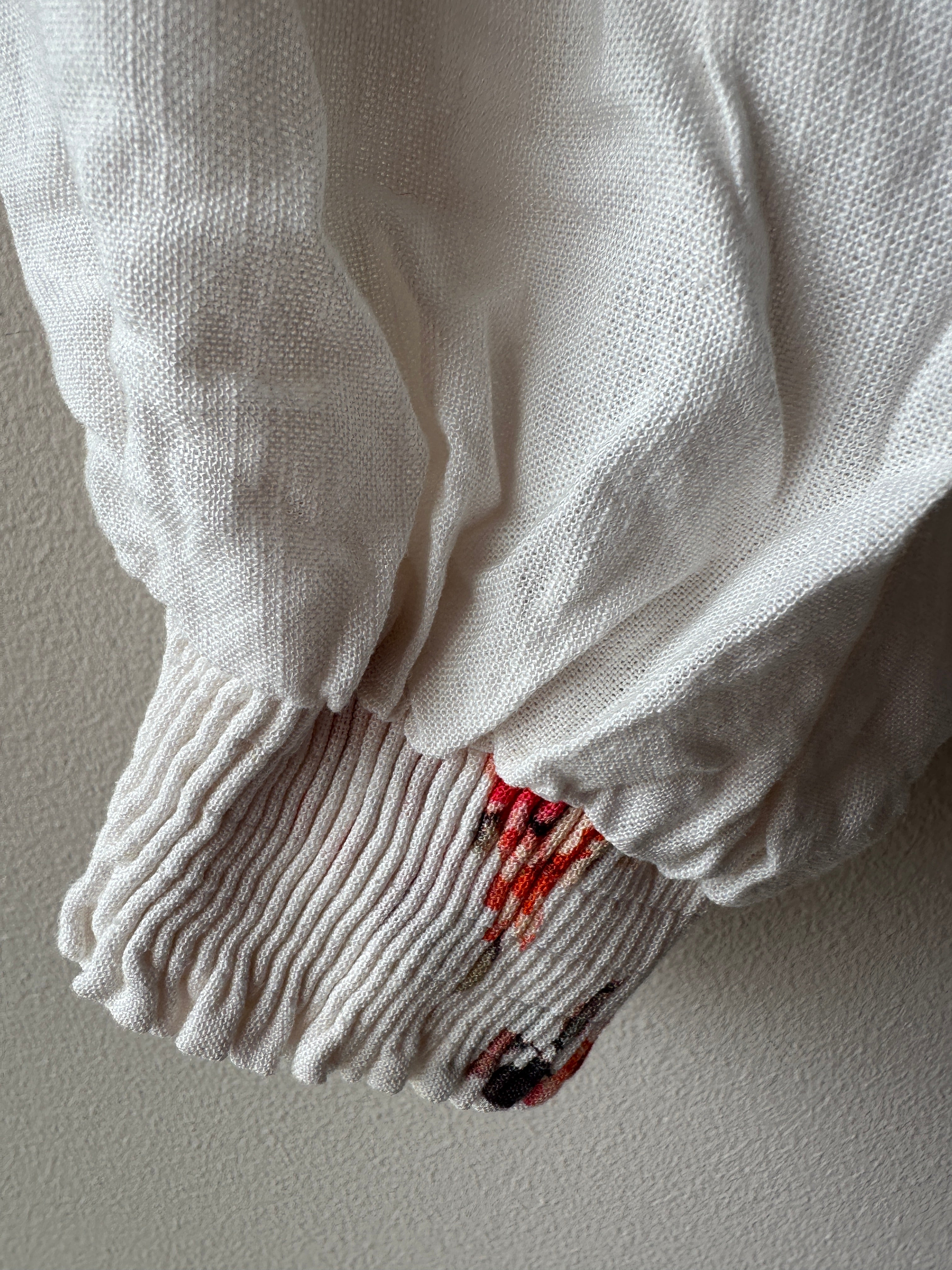 Närbild på smockad manschett på lång puffig ärm i vitt luftigt strukturerat tyg med inslag av rött blommigt tryck vid kanten.