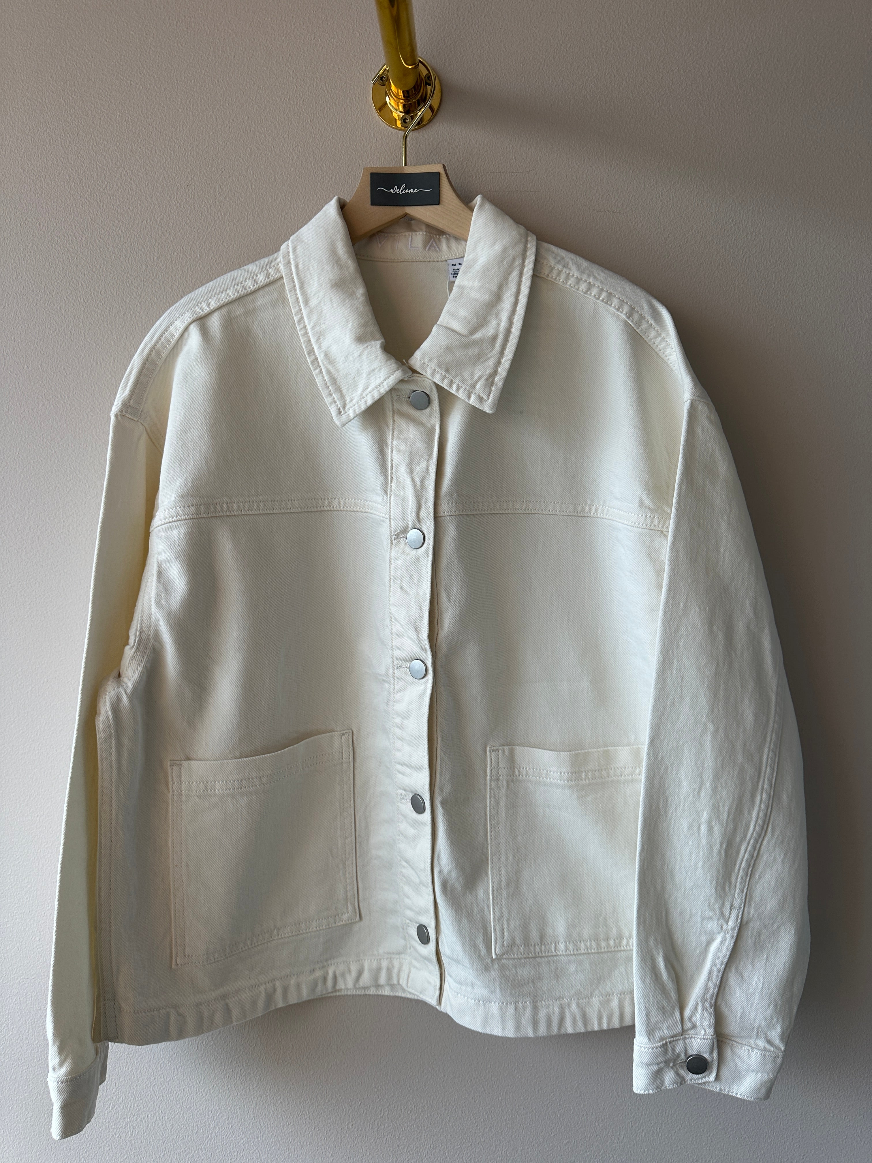 Vit oversized denimjacka med klassisk krage, tryckknappar och stora patchfickor, hängd på galge mot ljus vägg.