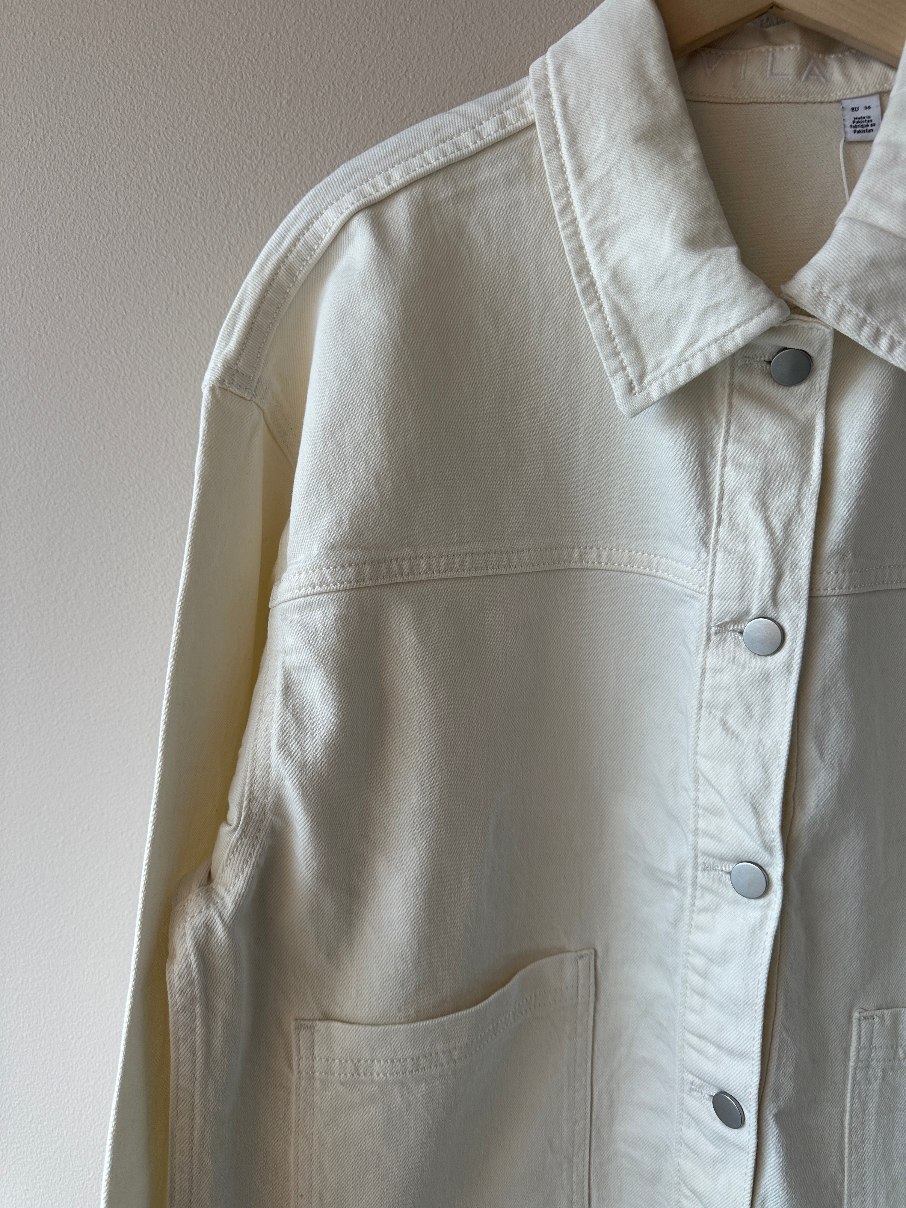 Närbild på överkropp av vit denimjacka med silvriga tryckknappar, klassisk krage och synliga sömdetaljer.