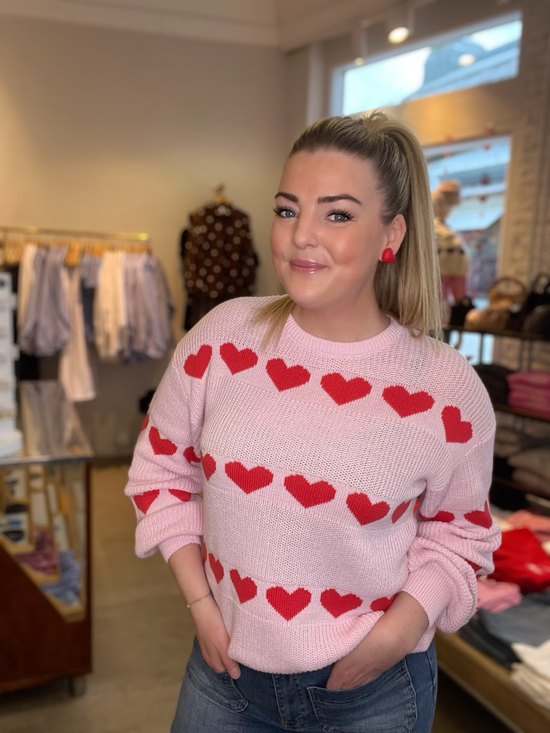 Leende kvinna med händerna i fickorna i pastellrosa stickad tröja med stora röda hjärtan i jacquardmönster, stylade med blå jeans och röda örhängen.