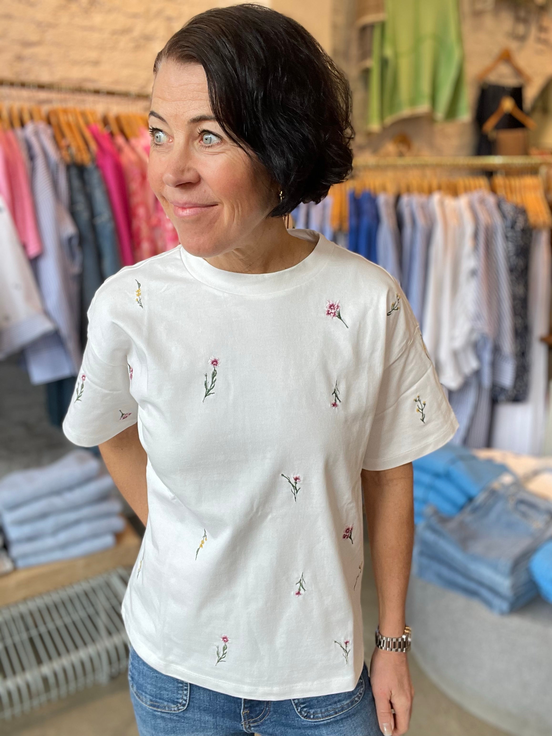 Kvinna i vit t-shirt med broderade småblommor i rosa och gult, rund hals och kortare ärm – stylade med blå jeans i butiksmiljö.