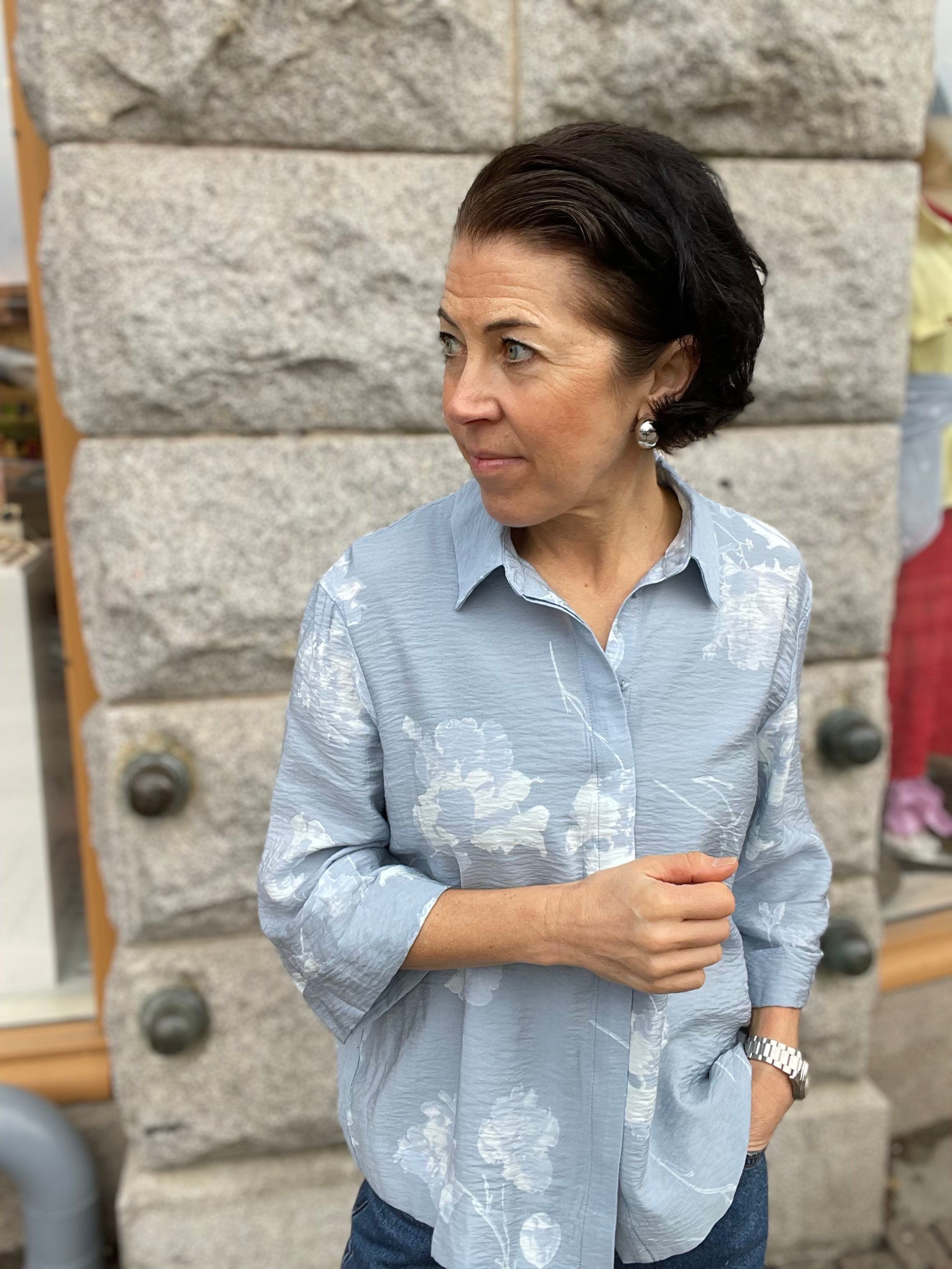 Kvinna med ena handen i fickan i ljusblå blommig jacquardskjorta med klassisk krage och 3/4-ärmar, stylade med blå jeans