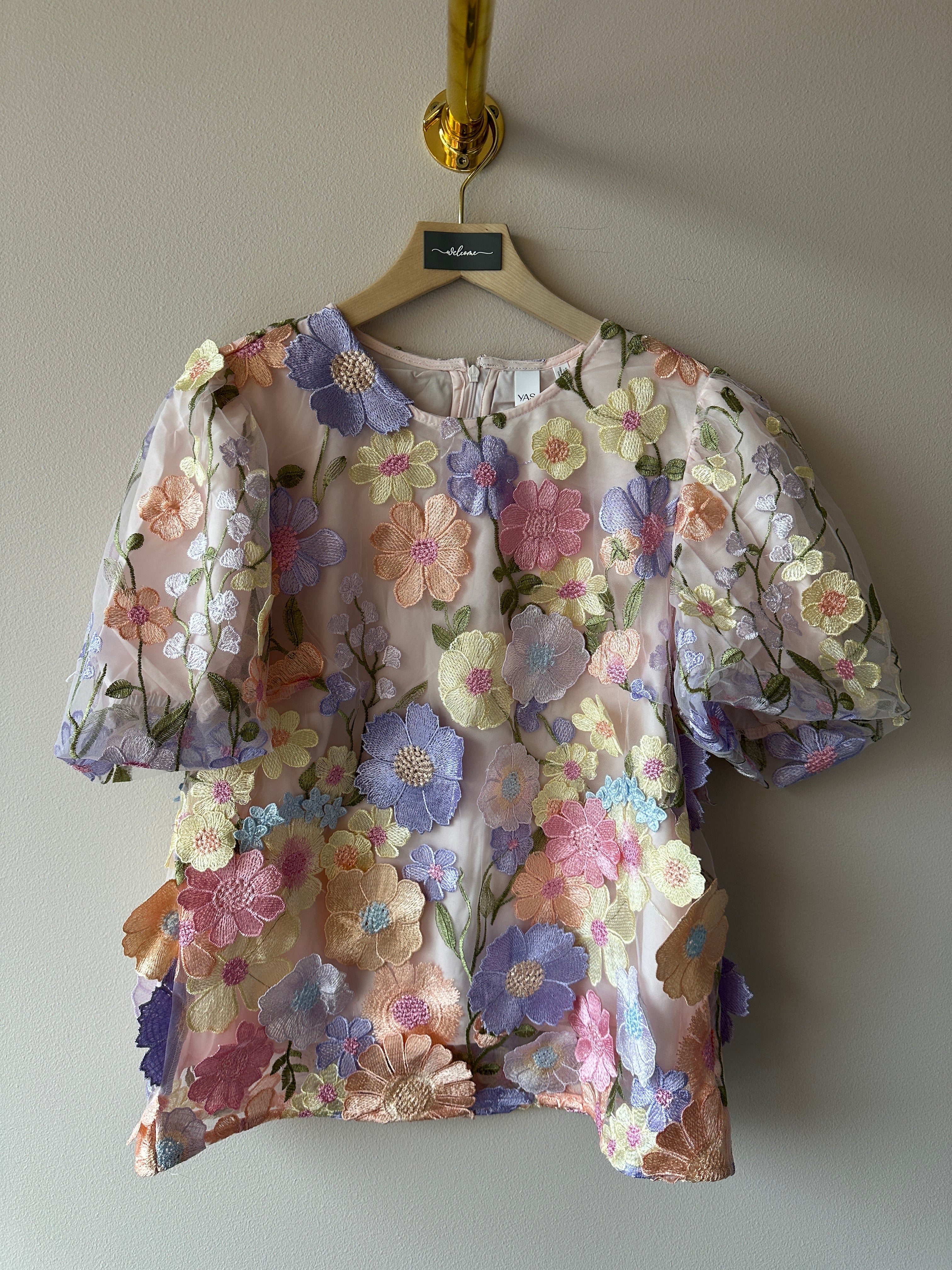 Lavendelfärgad topp med täta 3D-broderade pastellblommor i lila, rosa, gult och orange över hela tyget, rund hals med dragkedja bak och korta puffiga ärmar, hängande på galge mot beige vägg, etikett YAS synlig i halsen.
