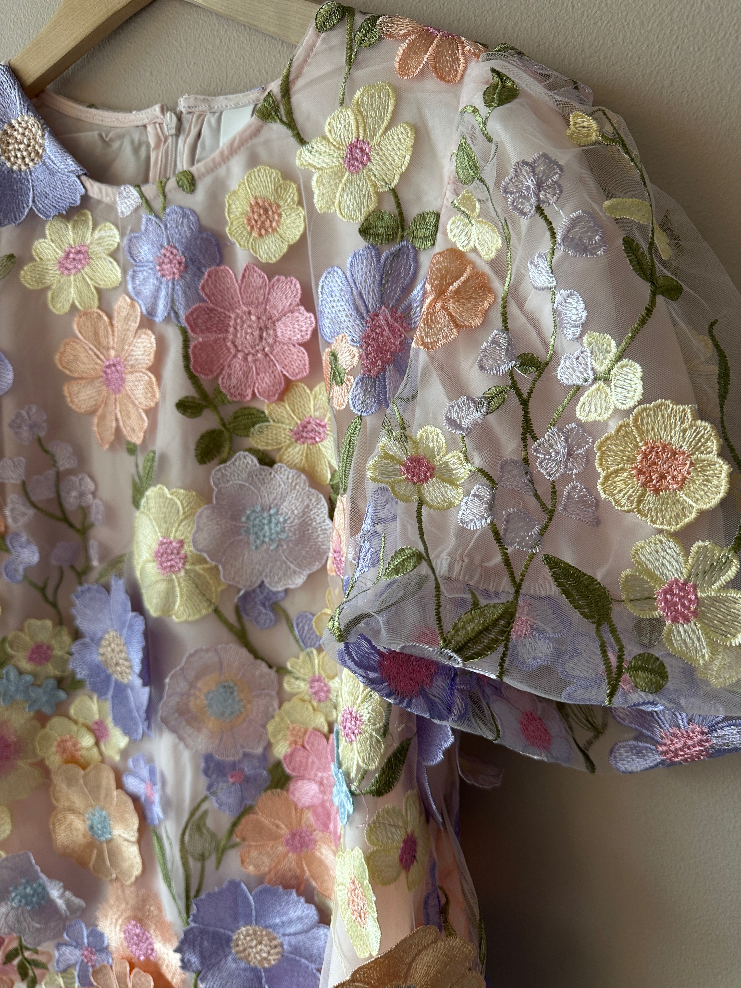 Närbild på övre del och puffig ärm av lavendelfärgad topp med täta 3D-broderade pastellblommor i flera färger på genomskinligt tyg, rund hals.